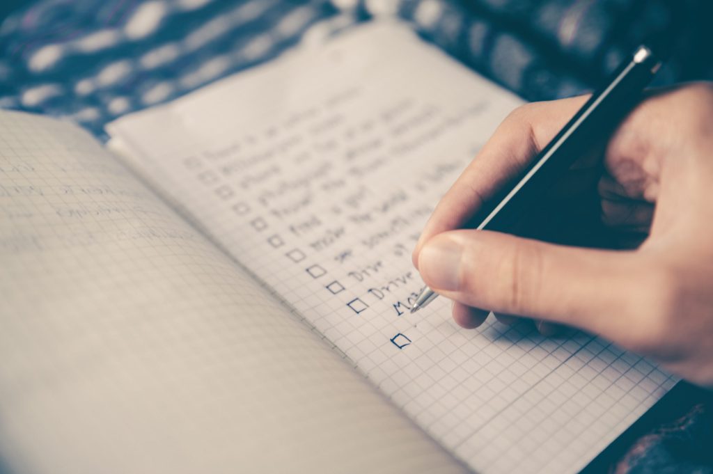 A hand holds a pen, writing a checklist in a notebook.